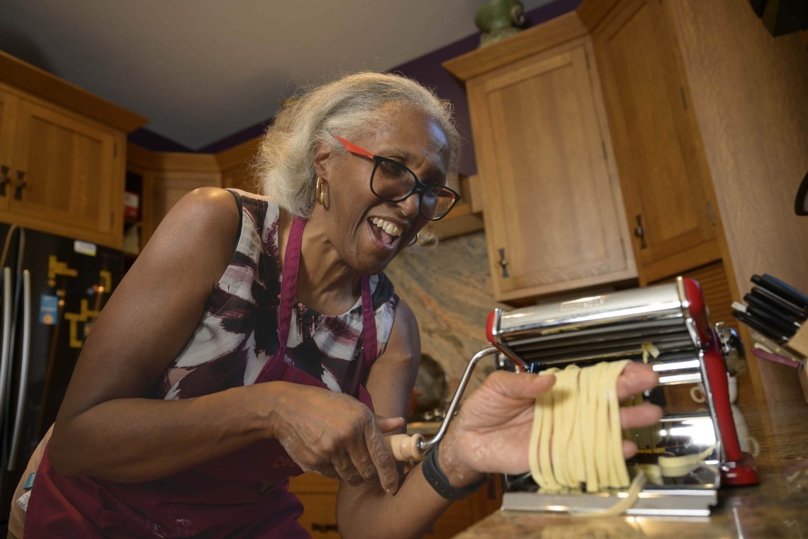 A person pulls linguine from a pasta-making machine. She has short gray hair and red glasses and is laughing.