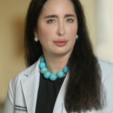 Person wearing a white lab coat and a turquoise necklace. She has long, straight dark hair.