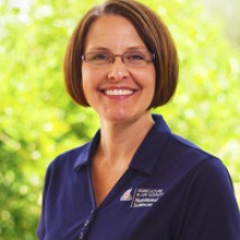 A person is smiling with short brown hair. She is wearing a blue polo shirt and rectangular glasses.